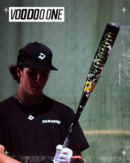 A baseball player in a black cap and shirt holds the 2027 DeMarini Voodoo One (-3) BBCOR Baseball Bat (WBD2705010), featuring an X14 Alloy Barrel and bold "VOODOO ONE" text on the bat's upper left.