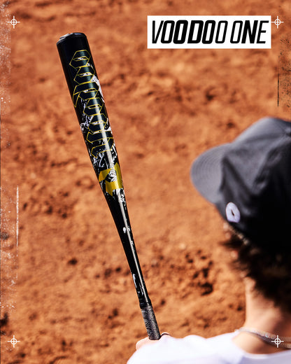 A person holds a black and yellow DeMarini 2027 Voodoo One (-3) BBCOR Baseball Bat (WBD2705010) over a dirt field, with "VOODOO ONE" text in the top right corner.