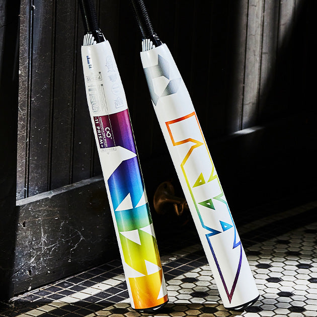 Two colorful umbrella bags with bold, geometric designs lean against a dark wooden door on a tiled floor. Each bag has a unique gradient color pattern and the word “MARY” printed in large letters.