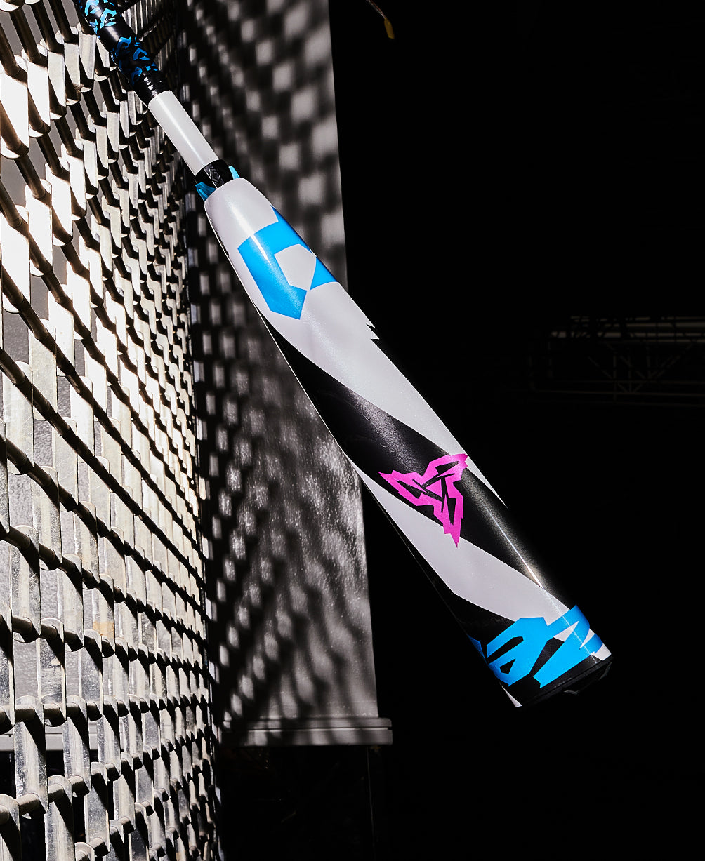 A 2025 DeMarini ZEN (-5) USSSA Baseball Bat (WBD2534010), white with blue and pink geometric designs, leans against a metal fence, casting a shadow in the bright sunlight.