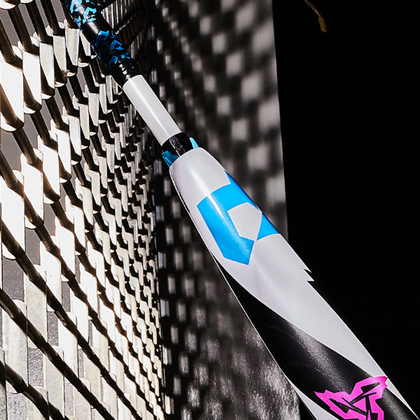 A close-up of a white baseball bat with blue and black geometric designs and a pink logo, leaning against a chain-link fence casting shadows.