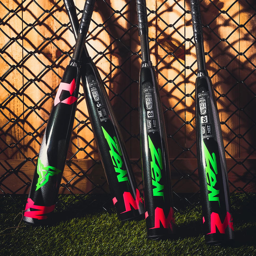 Four DeMarini ZEN Limited Edition (-10) USSSA baseball bats in neon green and pink, with composite barrels, lean against a chain-link fence on artificial turf as sunlight casts shadows on the wooden wall behind them.