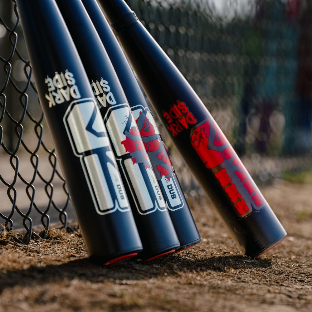Four black baseball bats lean against a chain-link fence on dirt, displaying Duke, Dark Side, and red graphics along their barrels—including the 2027 Easton Dub Darkside (-10) 2 3/4″ USSSA Baseball Bat from Easton, which features Double Barrel Construction and an Opti-Flex Handle for premium performance.