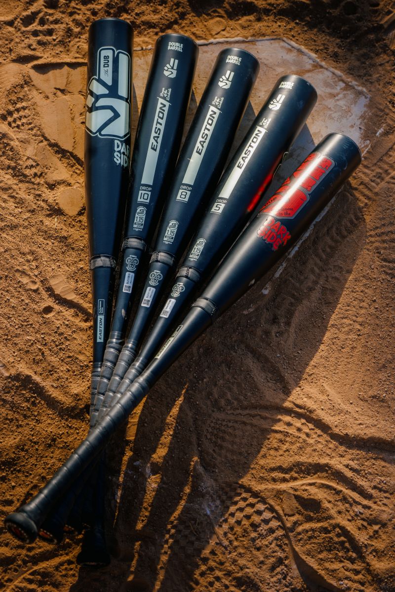 Five black baseball bats with assorted brand logos, including the 2027 Easton Dub Darkside (-10) 2 3/4″ USSSA Baseball Bat (EUT7DBDS10) by Easton, are neatly arranged side by side on sandy ground, with the handles positioned at the bottom and their barrels fanned out at the top.