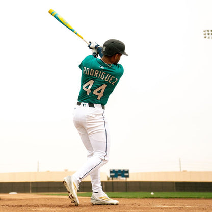 A baseball player in a green jersey with "Rodríguez" and 44 swings the 2025 Victus Vibe Pro-Crayon Julio Rodriguez (-5) USSSA bat by Victus, wearing a helmet and white pants. Overcast skies and buildings are visible in the background.