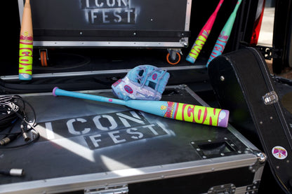A blue and purple glove and a colorful 2026 Rawlings Icon Fest (-10) USSSA Baseball Bat (RUT6IFL10) sit on a black ICON FEST case, with extra bats and a limited edition guitar case nearby.