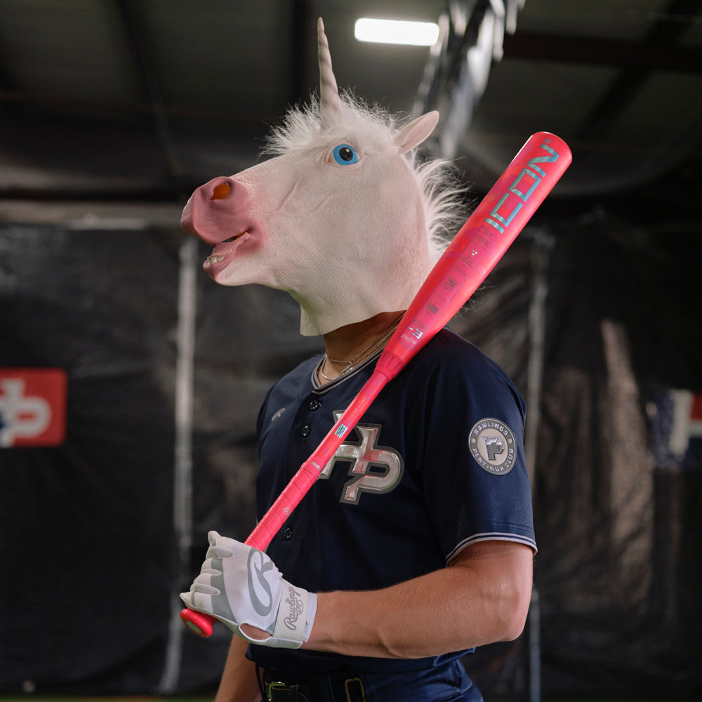 Inside an indoor sports facility, a person in a unicorn mask, navy baseball uniform, and white glove holds the Rawlings 2025 Icon Electric Unicorn (-3) BBCOR Baseball Bat (RBB5IPK3) in bright pink over their shoulder.
