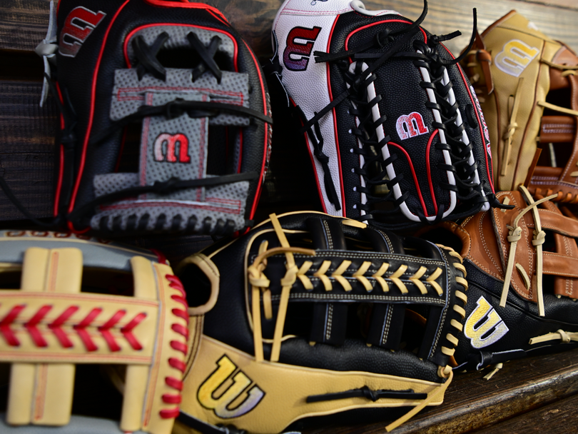 A close-up of five colorful baseball gloves, each with unique designs and stitching, arranged on a wooden surface. The gloves display prominent logos and a variety of colors including black, tan, red, and white.