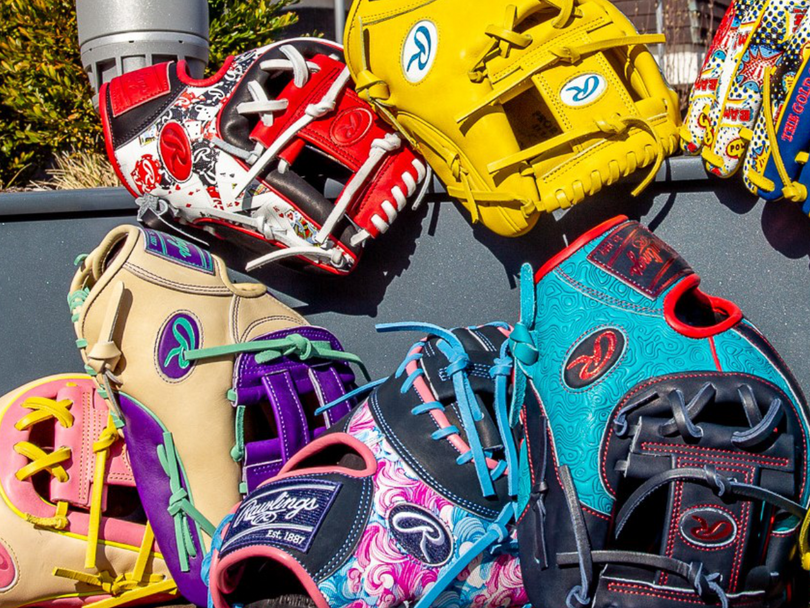 A collection of colorful and uniquely designed baseball gloves are displayed together outdoors, featuring various patterns, logos, and vibrant colors such as yellow, pink, blue, and red.