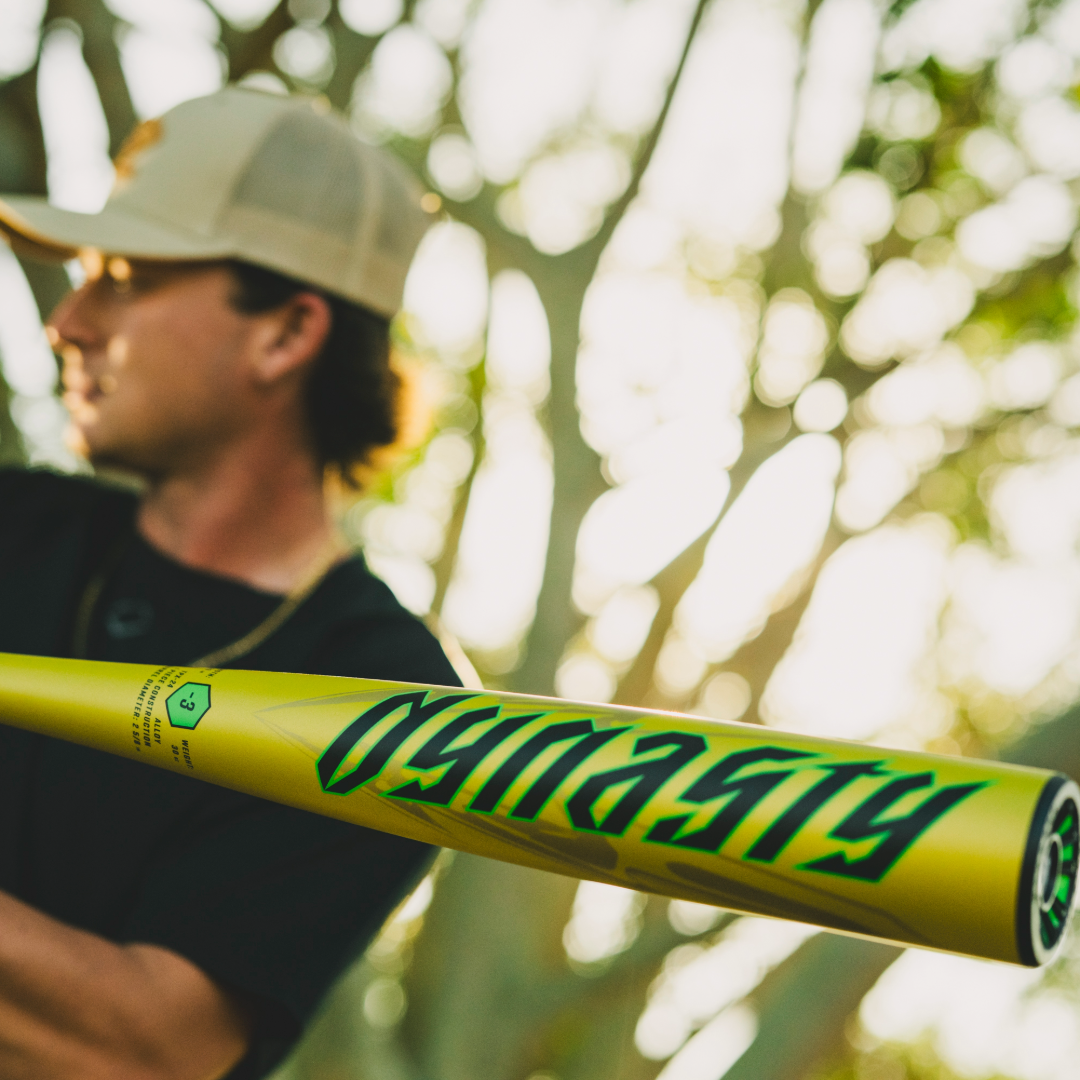 A person in a tan cap holds the 2026 Louisville Slugger Dynasty (-3) BBCOR Baseball Bat (WBL4162010), featuring DYNASTY in bold green letters, sharply focused against a blurred outdoor background.