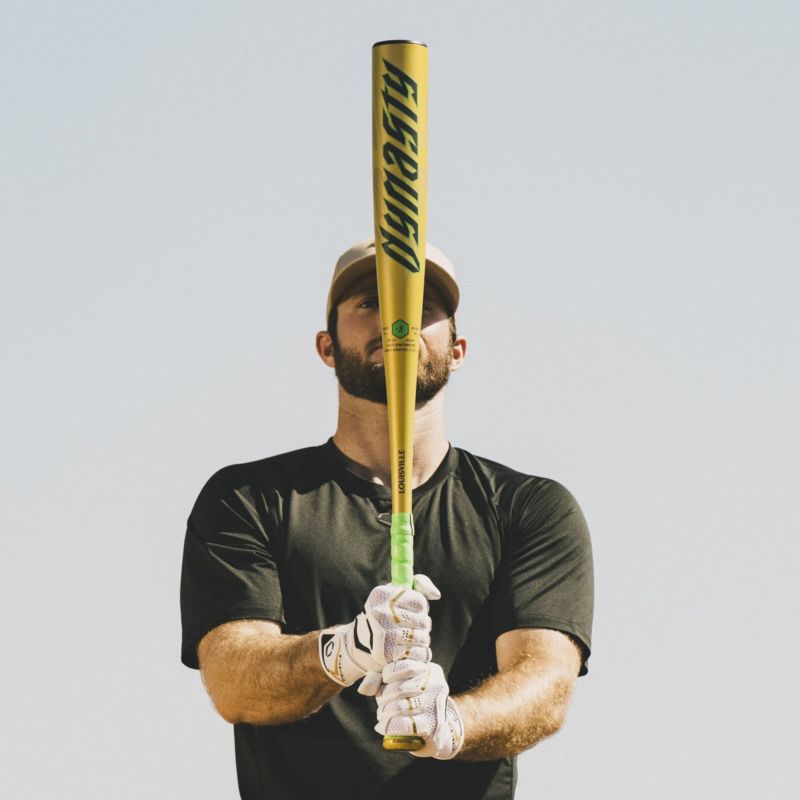 A man in a cap and black shirt, wearing white gloves, holds the 2026 Louisville Slugger Dynasty (-3) BBCOR Baseball Bat (WBL4162010) upright in front of his face against a clear sky.