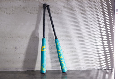 Two 2025 DeMarini The Goods Teal (-3) BBCOR Baseball Bats (WBD2564010) with black handles and yellow graphics lean against a concrete wall, their X14 Alloy Barrels standing out in the light.
