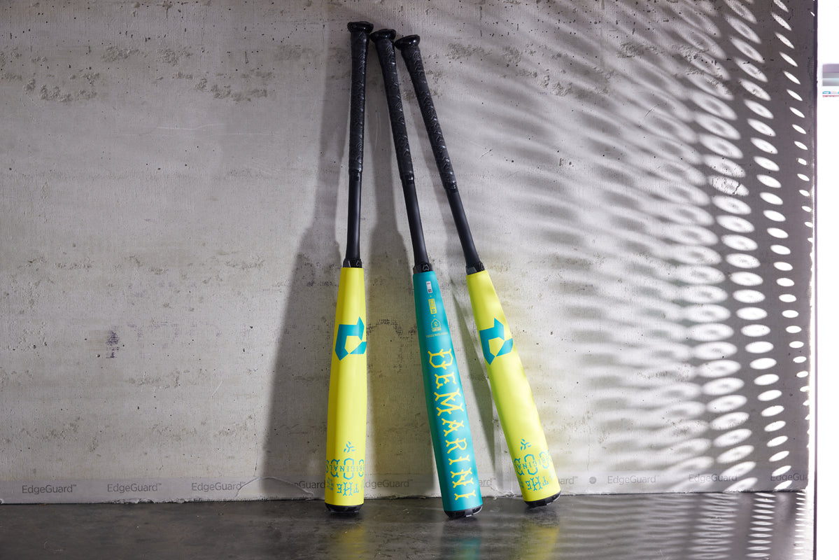Three 2025 DeMarini The Goods Neon Yellow (-3) BBCOR Baseball Bats (WBD2563010) with teal branding and X14 Alloy Barrel lean against a concrete wall, highlighted by patterned shadows and sunlight.