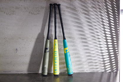 Three vibrant bats—gray, yellow, and teal—stand upright by a sunlit concrete wall, showcasing bold colors inspired by the 2025 DeMarini The Goods Teal (-3) BBCOR Baseball Bat: WBD2564010 from DeMarini.