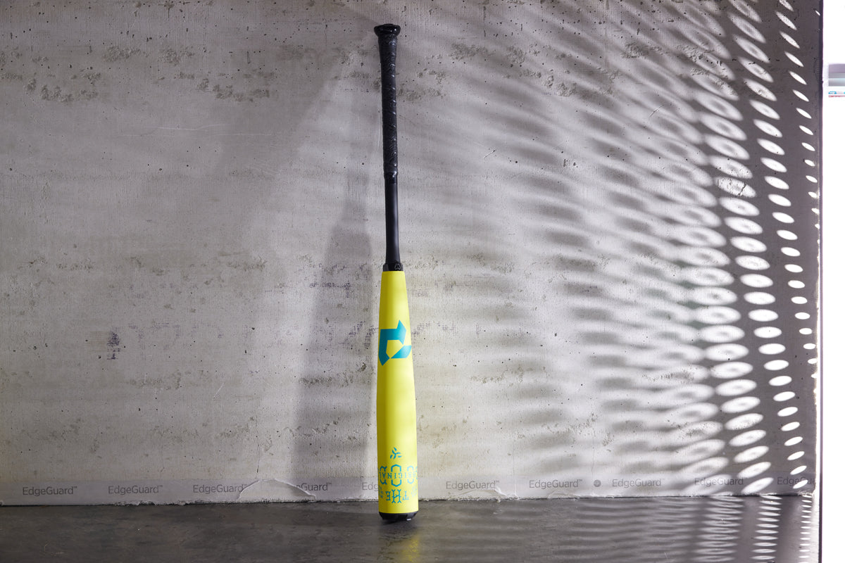 A 2025 DeMarini The Goods Neon Yellow (-3) BBCOR Baseball Bat (WBD2563010) stands upright against a concrete wall, blue accents and patterns of light and shadow highlighting its design.