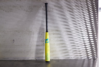 A 2025 DeMarini The Goods Neon Yellow (-3) BBCOR Baseball Bat (WBD2563010) stands upright against a concrete wall, blue accents and patterns of light and shadow highlighting its design.