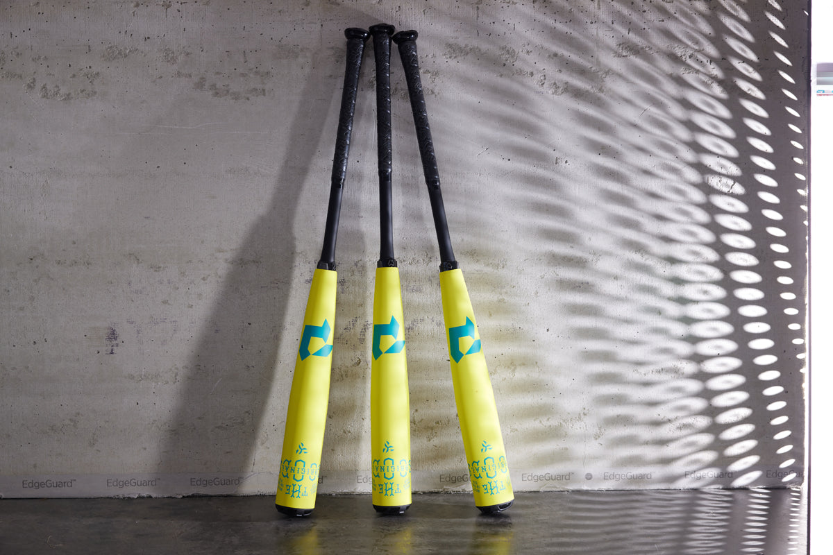 Three yellow and black DeMarini baseball bats, including the 2025 The Goods Neon Yellow (-3) BBCOR Bat (WBD2563010), stand upright against a concrete wall, their handles touching and casting patterned shadows on the surface.