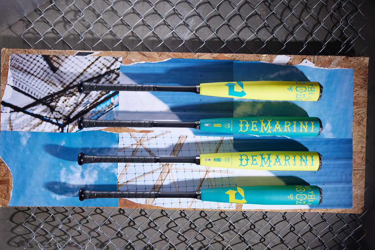 Three teal 2025 DeMarini The Goods (-3) BBCOR Baseball Bats (WBD2564010) are displayed on a board featuring a sky and chain-link fence print, set against an actual chain-link fence in the background.