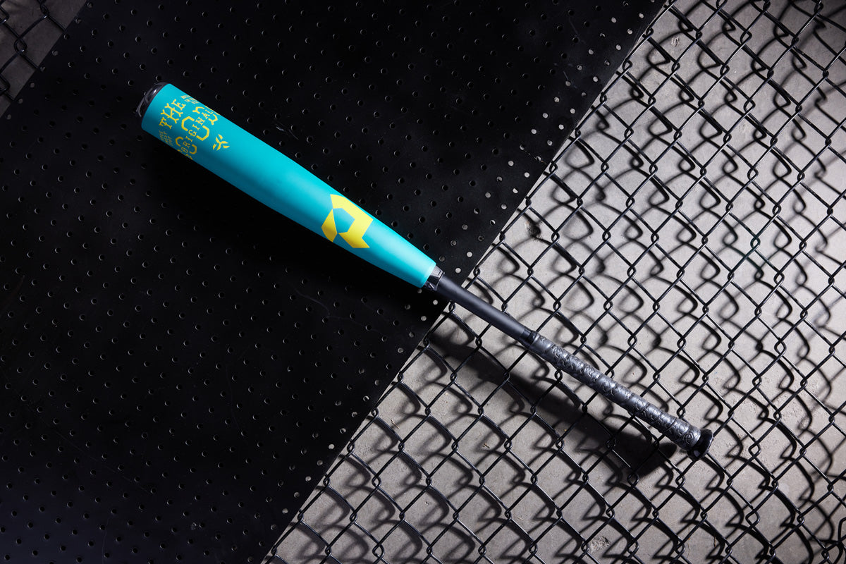 A teal and yellow 2025 DeMarini The Goods Teal (-3) BBCOR Baseball Bat (WBD2564010) with a black grip rests diagonally on a surface split between perforated black metal and chain-link fencing.