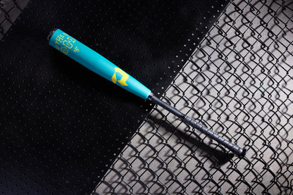 A teal and yellow 2025 DeMarini The Goods Teal (-3) BBCOR Baseball Bat (WBD2564010) with a black grip rests diagonally on a surface split between perforated black metal and chain-link fencing.