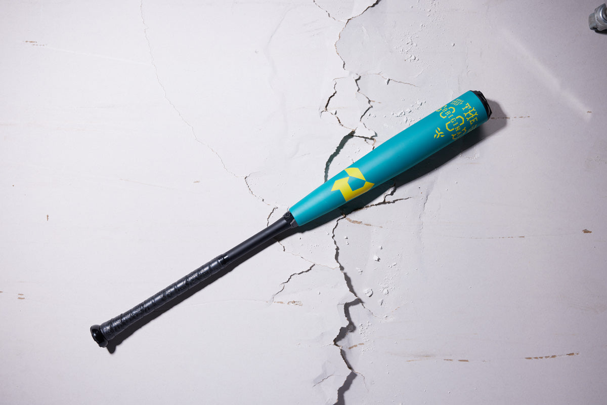 A 2025 DeMarini The Goods Teal (-3) BBCOR Baseball Bat: WBD2564010 rests on a cracked white concrete surface. The teal and black bat features yellow graphics and THE GOODS branding on its X14 Alloy Barrel.