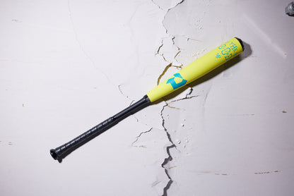 A 2025 DeMarini The Goods Neon Yellow (-3) BBCOR Baseball Bat (WBD2563010) in yellow and black rests on cracked white concrete, casting a shadow to the right.