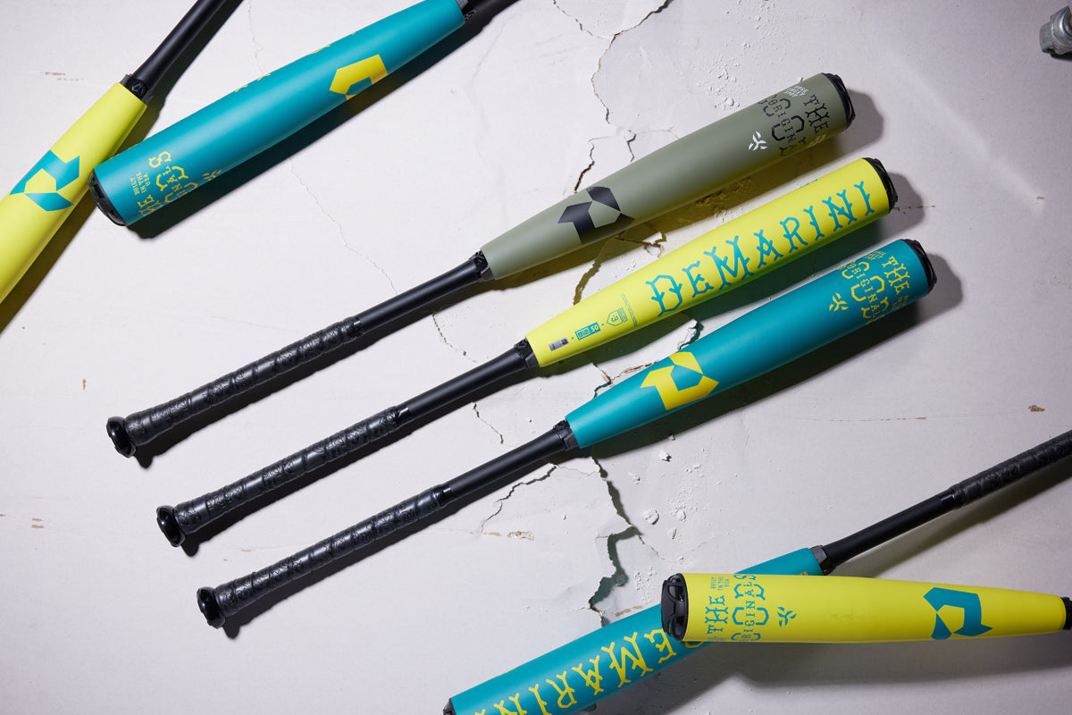 Four 2025 DeMarini The Goods Teal (-3) BBCOR Baseball Bats (WBD2564010) with black handles are placed diagonally on a cracked white surface, displaying vibrant teal, yellow, and green X14 Alloy Barrel designs for top hybrid performance.