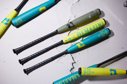 Four 2025 DeMarini The Goods Teal (-3) BBCOR Baseball Bats (WBD2564010) with black handles are placed diagonally on a cracked white surface, displaying vibrant teal, yellow, and green X14 Alloy Barrel designs for top hybrid performance.