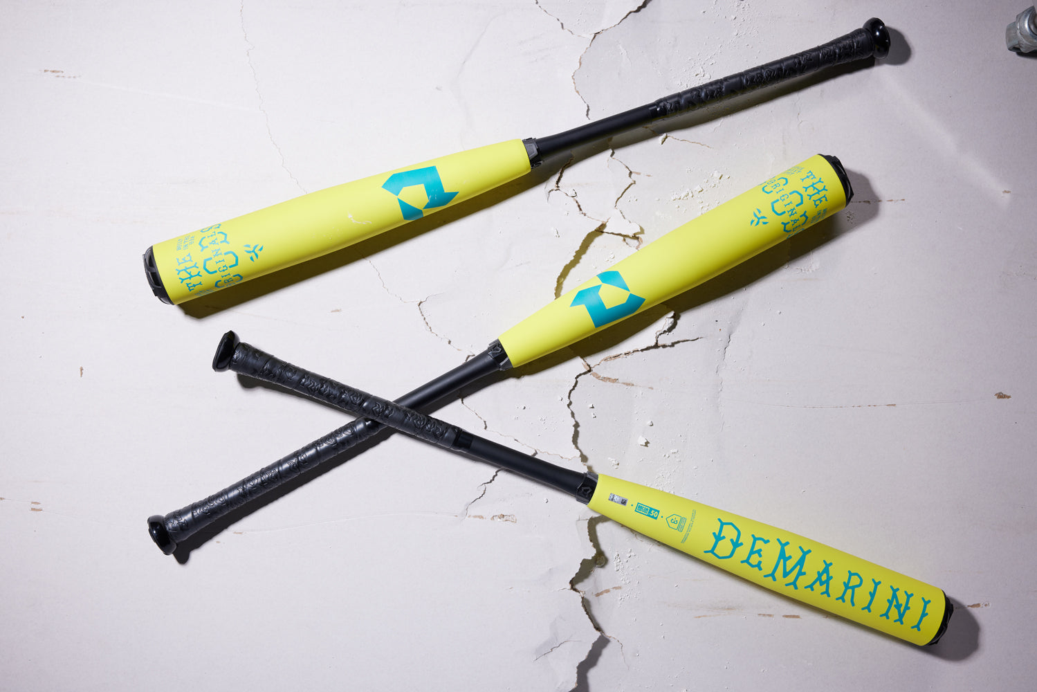 Three yellow DeMarini baseball bats with black handles and blue designs, including the 2025 DeMarini The Goods Neon Yellow (-3) BBCOR Baseball Bat (WBD2563010), lie crossed on a cracked white surface.