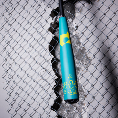 A DeMarini 2025 The Goods Teal (-3) BBCOR Baseball Bat: WBD2564010 hangs in front of a chain-link fence with a large hole and broken concrete, highlighting the bat's power and performance.