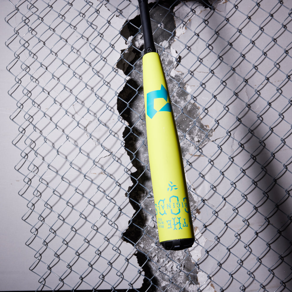 A 2025 DeMarini The Goods Neon Yellow (-3) BBCOR Baseball Bat hangs vertically with blue accents, appearing to smash through a chain-link fence and concrete—highlighting the bold power of its X14 Alloy Barrel.