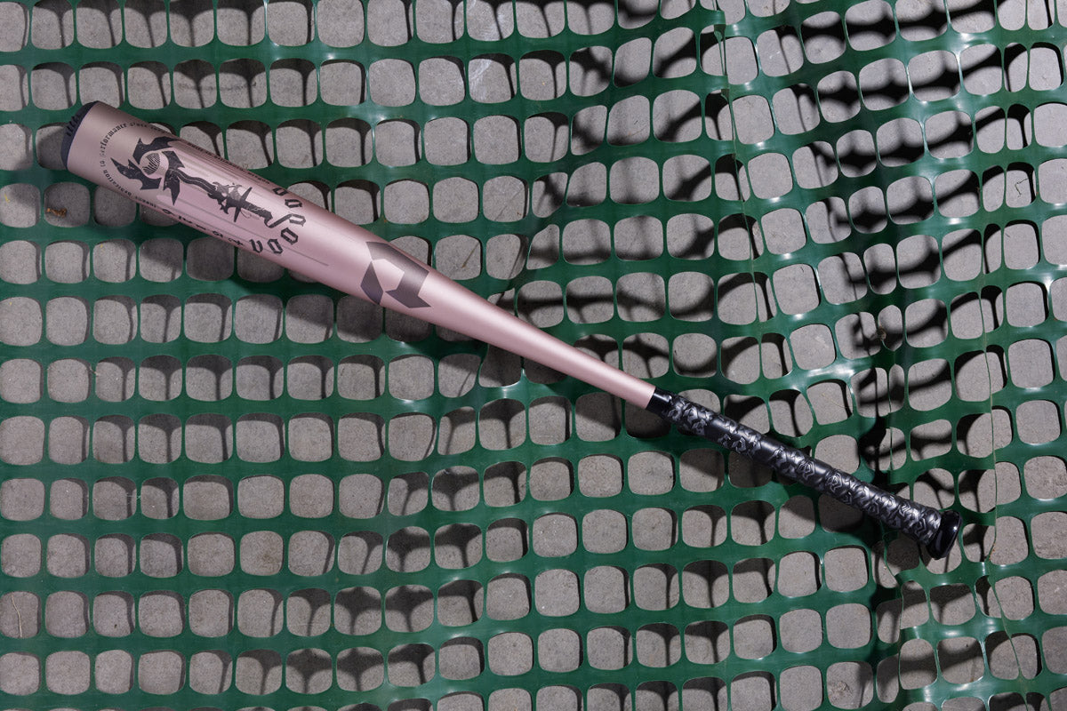 The 2025 DeMarini Voodoo One Rose Gold (-3) BBCOR Baseball Bat (WBD2628010) with a black grip rests diagonally on green mesh fencing, highlighting its sleek X14 Alloy Barrel design.