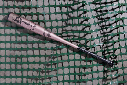 The 2025 DeMarini Voodoo One Rose Gold (-3) BBCOR Baseball Bat (WBD2628010) with a black grip rests diagonally on green mesh fencing, highlighting its sleek X14 Alloy Barrel design.