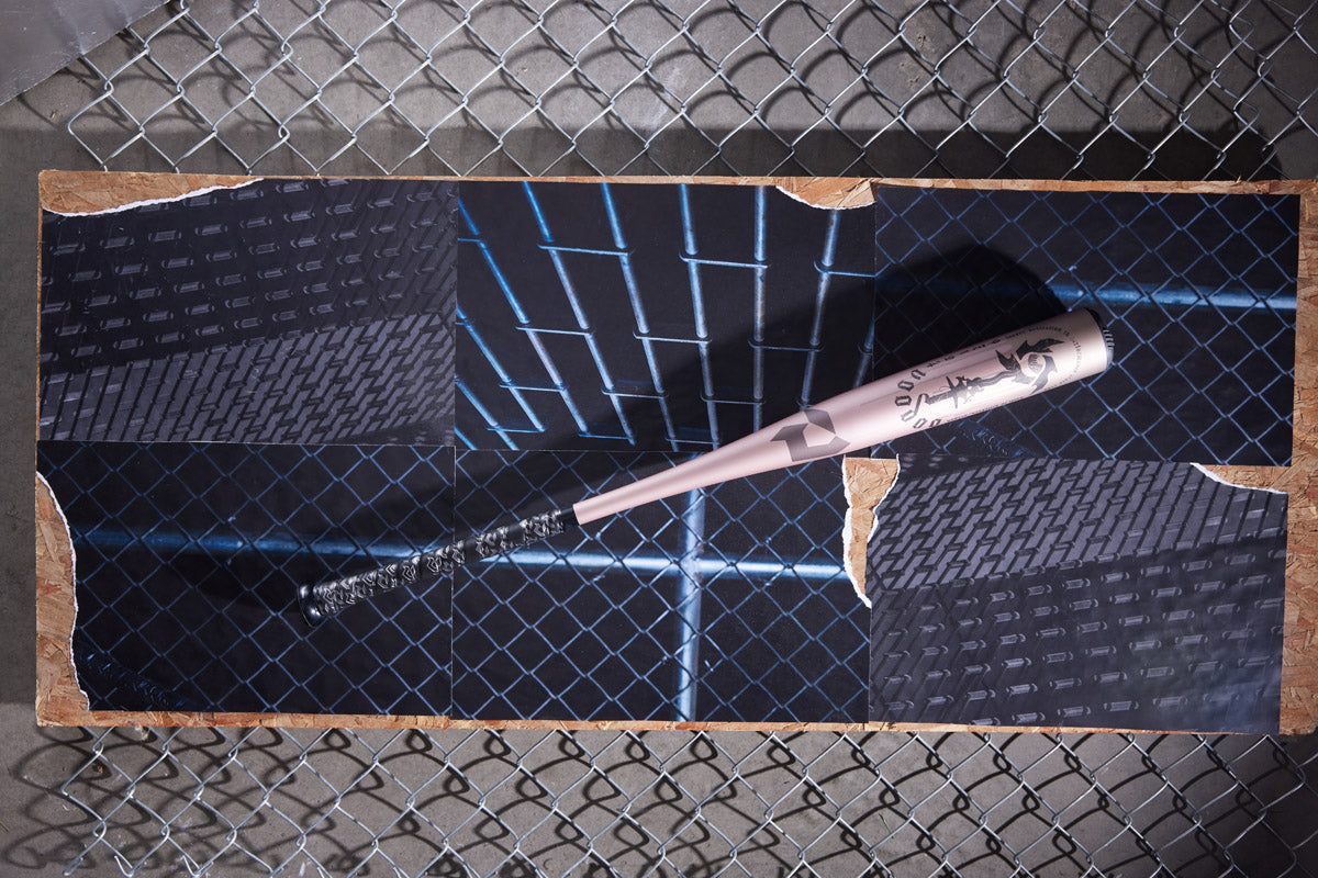 A pink eyeliner pen lies diagonally on vibrant photos of blue-lit chain-link fences and brick walls, set atop a wooden board—echoing the sleek, modern design of the DeMarini 2025 Voodoo One Rose Gold (-3) BBCOR Baseball Bat: WBD2628010.