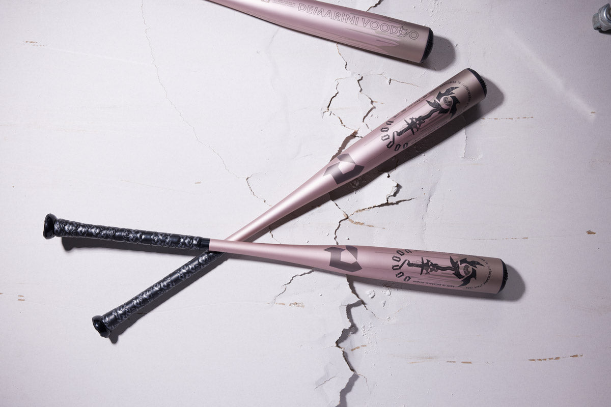 Two 2025 DeMarini Voodoo One Rose Gold (-3) BBCOR Baseball Bats with black grips and artwork are crossed on a cracked, light surface; a third DeMarini bat is partially visible at the top.