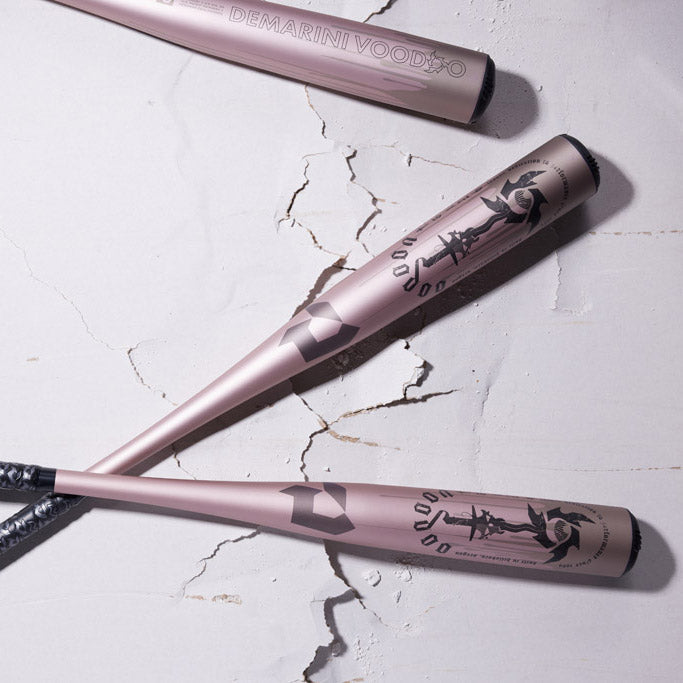 Three pink DeMarini Voodoo baseball bats with black graphics rest on a cracked white surface, arranged diagonally with two bats crossing each other near the handles.