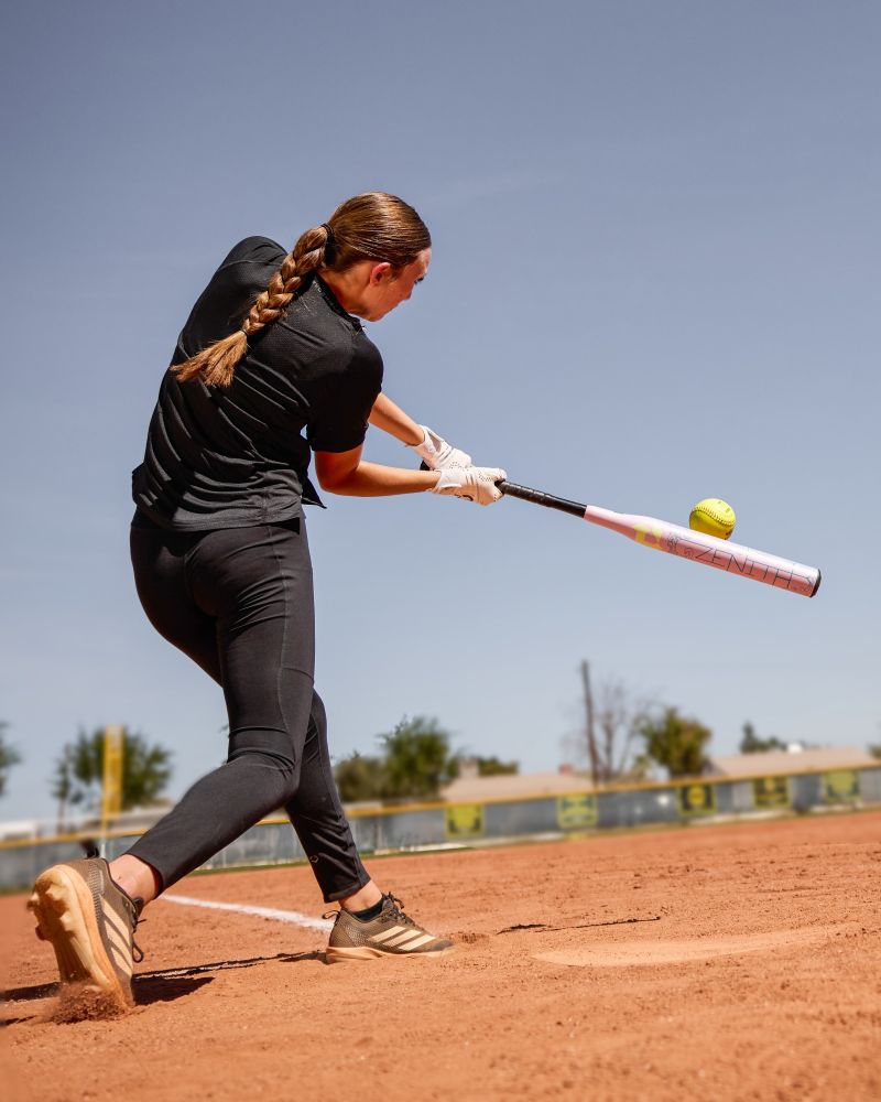 A softball player in black athletic wear swings the 2027 DeMarini Zenith (-13) Fastpitch Softball Bat (WBD2732010), connecting with a yellow softball on a dirt field beneath a clear blue sky.