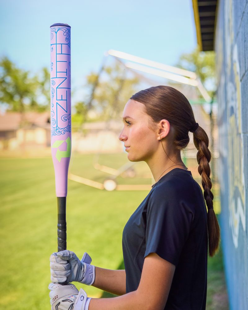 A young woman with a long braid stands on a grassy field, wearing a black shirt and white gloves, holding the DeMarini 2027 Zenith (-13) Fastpitch Softball Bat (WBD2732010), facing sideways with a focused gaze.