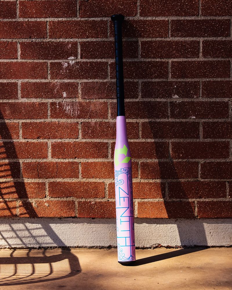A 2027 DeMarini Zenith (-13) Fastpitch Softball Bat (WBD2732010), featuring purple, pink, and blue colors and "ZENITH" branding, stands upright against a red brick wall with a fence shadow nearby.