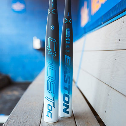 Two Easton Ghost OG (-11) Fastpitch Softball Bats by Easton stand upright near wooden dugout benches, featuring Double Barrel construction, with a blurred blue wall in the background.