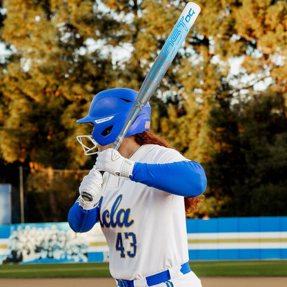 A UCLA softball player in a blue helmet stands ready to bat, holding an Easton 2025 Ghost OG (-11) Fastpitch Softball Bat (EFP5GHOG11) over their shoulder on a field with trees in the background.