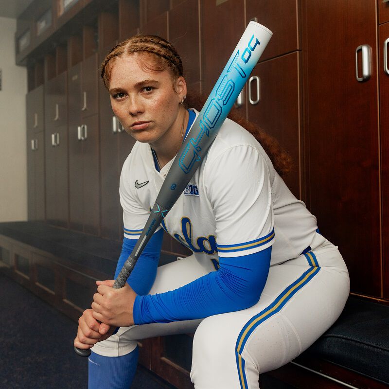 A softball player in a white and blue uniform sits in the locker room, confidently holding her 2025 Easton Ghost OG (-11) Fastpitch Softball Bat: EFP5GHOG11, with braided hair and blue sleeves under her jersey.