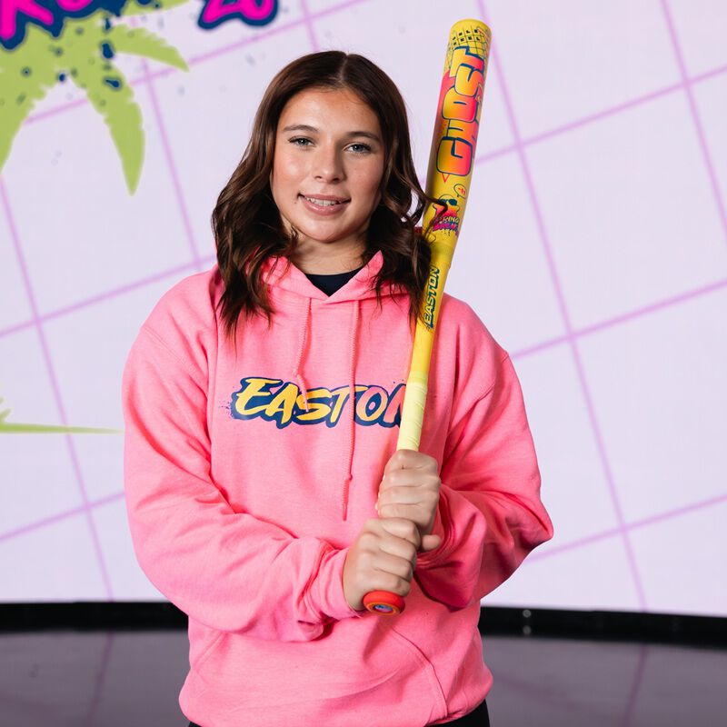 A young woman in a bright pink Easton hoodie holds the 2026 Easton Ghost Advanced Spring Break Yellow (-10) Fastpitch Softball Bat (EFP6GHSY10), standing before a retro, colorful backdrop with palm tree and diagonal line graphics.