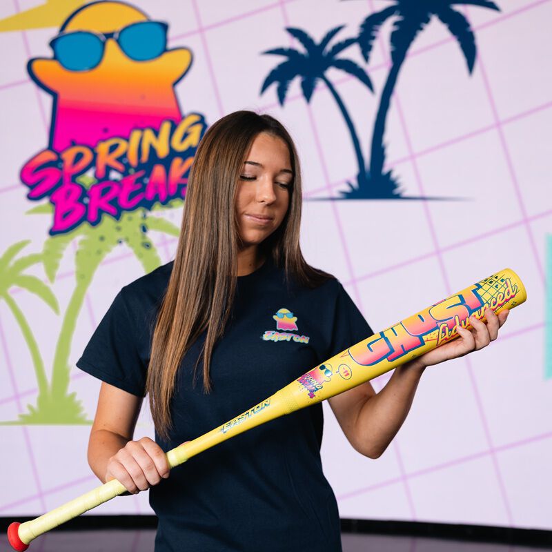 A woman with long brown hair in a navy t-shirt holds the 2026 Easton Ghost Advanced Spring Break Yellow (-11) Fastpitch Softball Bat (EFP6GHSY11) in front of a lively Spring Break sign featuring palm trees and a sun with sunglasses.