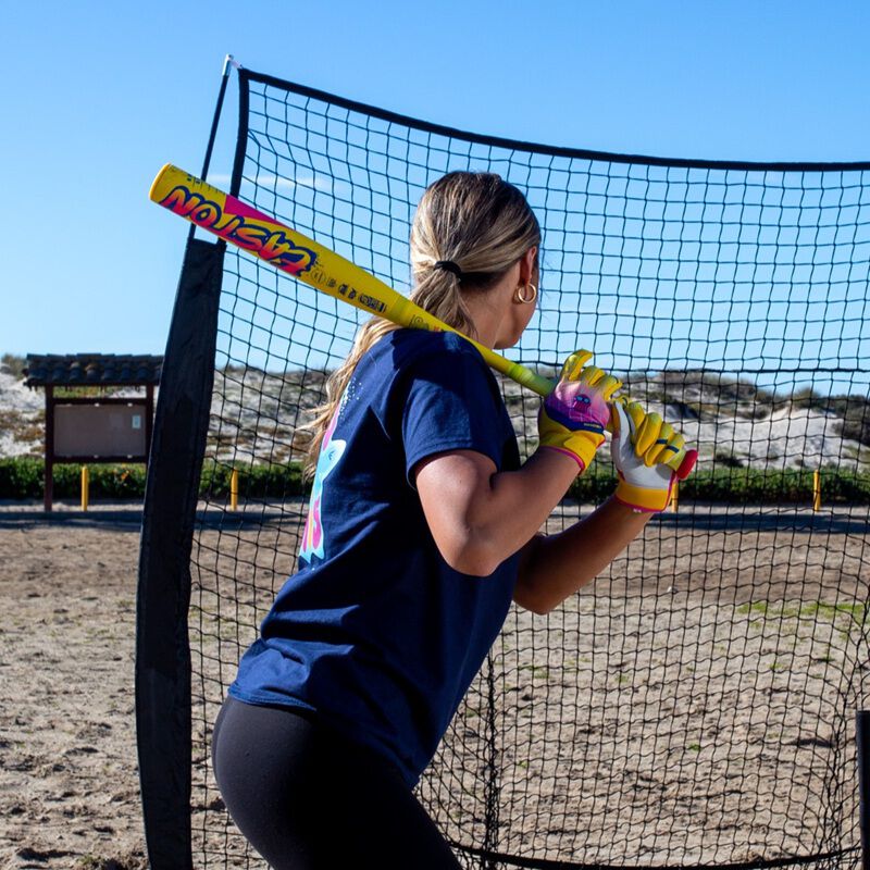 A woman in a navy shirt and yellow gloves holds the 2026 Easton Ghost Advanced Spring Break Yellow (-10) Fastpitch Softball Bat (EFP6GHSY10), standing ready to bat outdoors on sandy ground beneath a blue sky.