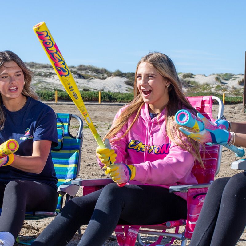 Three young women sit on sandy dunes under a blue sky, smiling and holding bright yellow 2026 Easton Ghost Advanced Spring Break (-10) Fastpitch Softball Bats (EFP6GHSY10) from Easton.