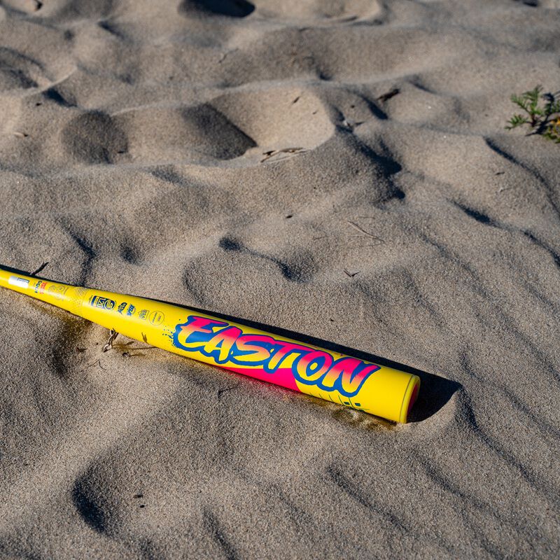 A 2026 Easton Ghost Advanced Spring Break Yellow (-10) Fastpitch Softball Bat with vibrant branding and Sonic Comp Max composite sits partly buried in light brown sand, small plants in the background.