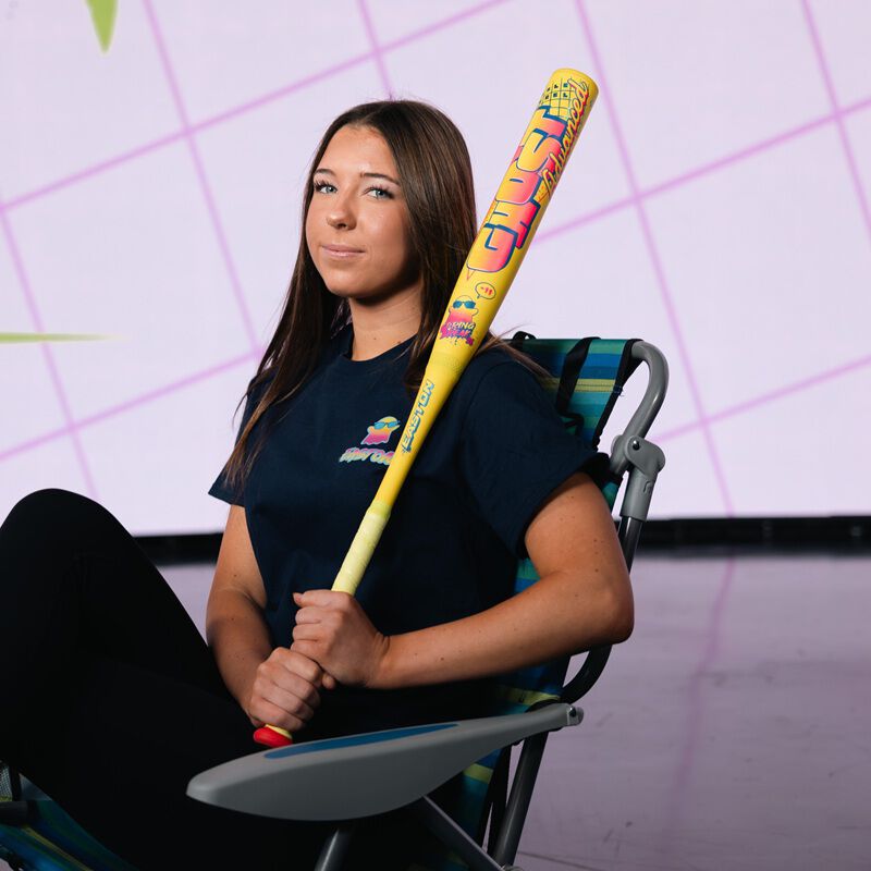 A young woman in black sits indoors on a colorful lawn chair, confidently holding the 2026 Easton Ghost Advanced Spring Break Yellow (-10) Fastpitch Softball Bat (EFP6GHSY10) by Easton, against a light grid-patterned background.