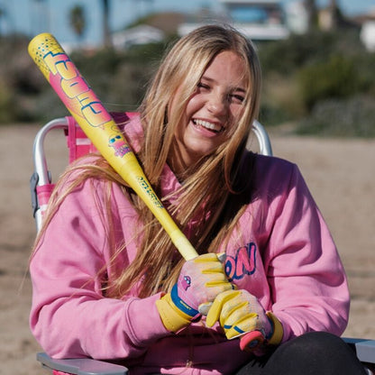 A smiling young woman with long blonde hair sits outside in a pink chair, wearing a pink hoodie and yellow gloves, holding the 2026 Easton Ghost Advanced Spring Break Yellow (-10) Fastpitch Softball Bat by Easton.