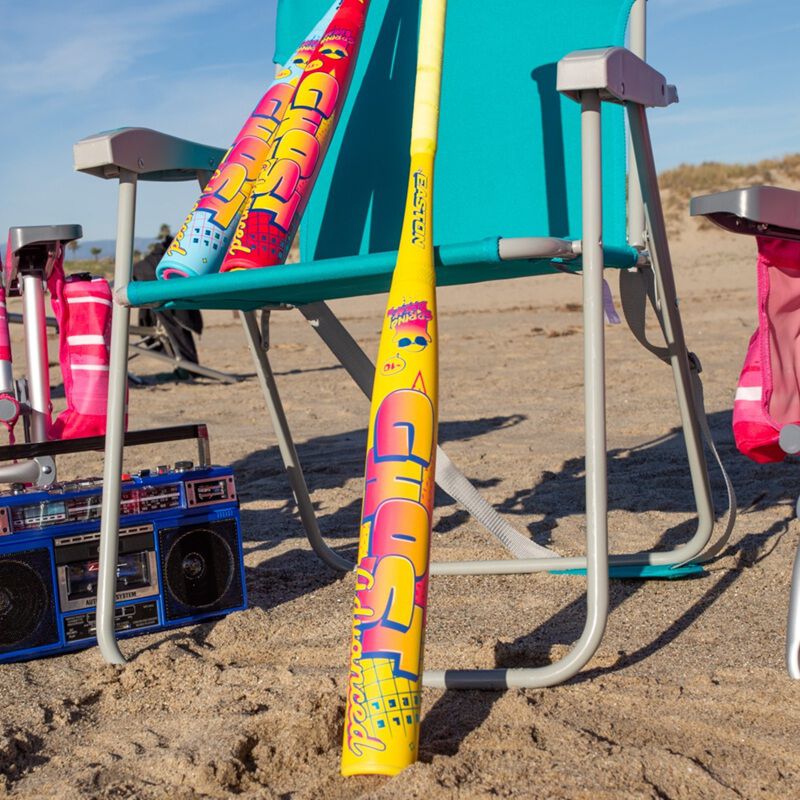 A 2026 Easton Ghost Advanced Spring Break Yellow (-10) Fastpitch Softball Bat (EFP6GHSY10) by Easton, featuring a vibrant design, leans against a turquoise beach chair on sand with another bat, a boom box, and bags nearby.